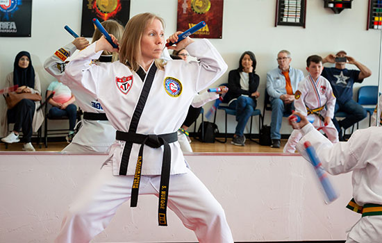 An adult black belt practices with nunchucks while a student trains beside her.