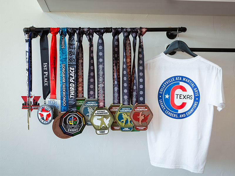 Songahm Taekwondo medals on a rack beside a Colleyville ATA Texas shirt.