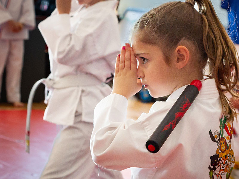 A preschool student in a white uniform holds a practice nunchuck at her shoulder during class.